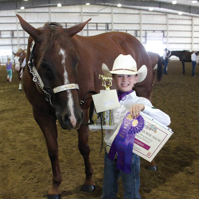 A small girl wearing a cowboy hat proudly holds up a purple ribbon while standing next to her horse.